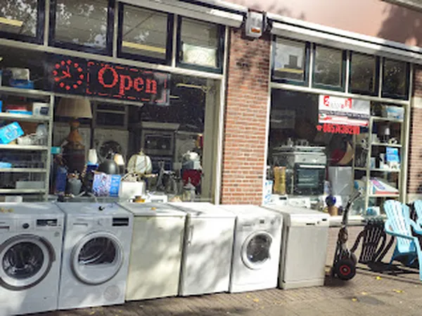 Voorgevel van kringloopwinkel met wasmachiines en drogers uitgestald voor het raam, neonreclame 'Open' zichtbaar