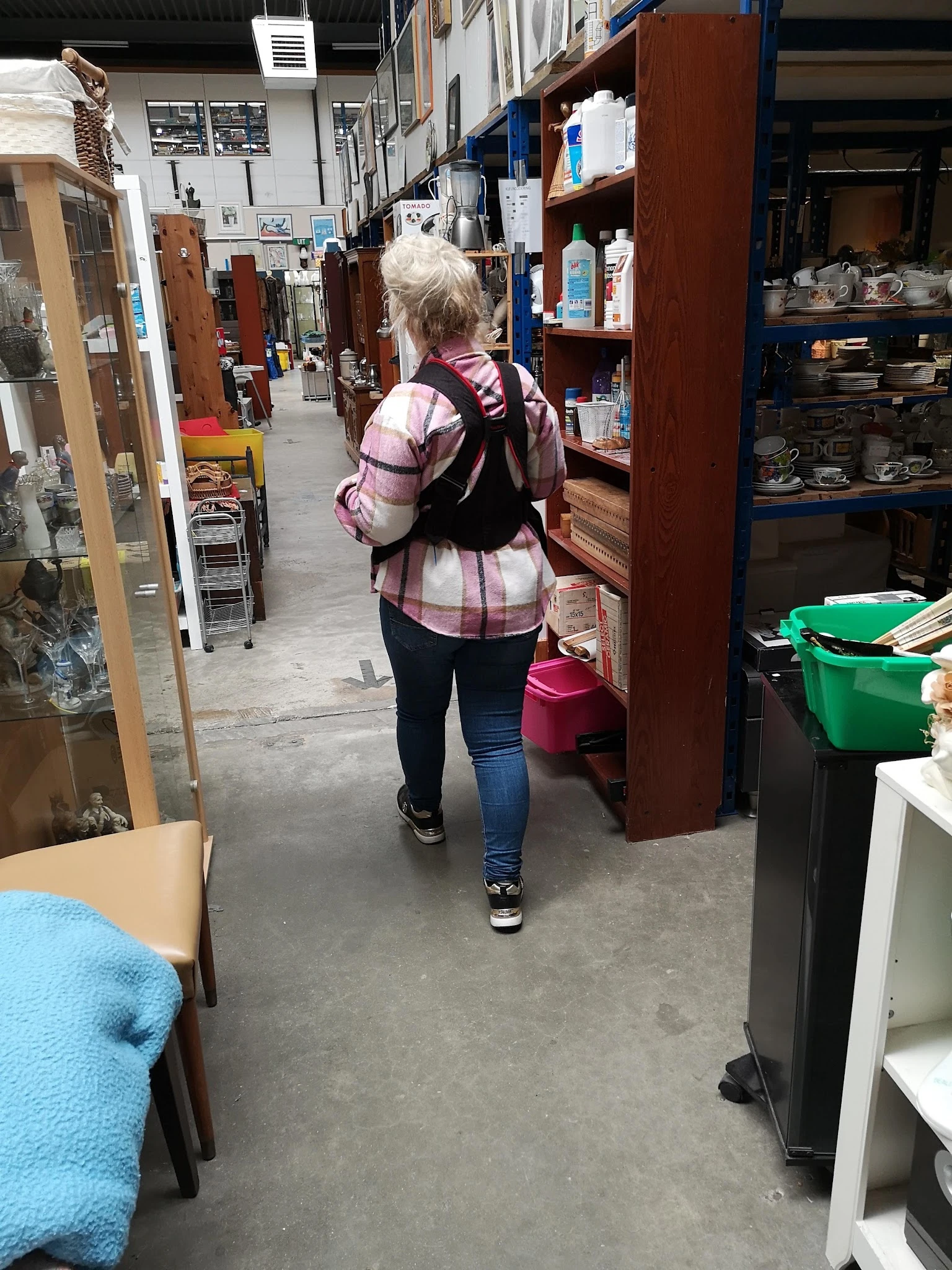 Persoon loopt door winkelpad tussen stellingen met tweedehands goederen en groene kratjes.