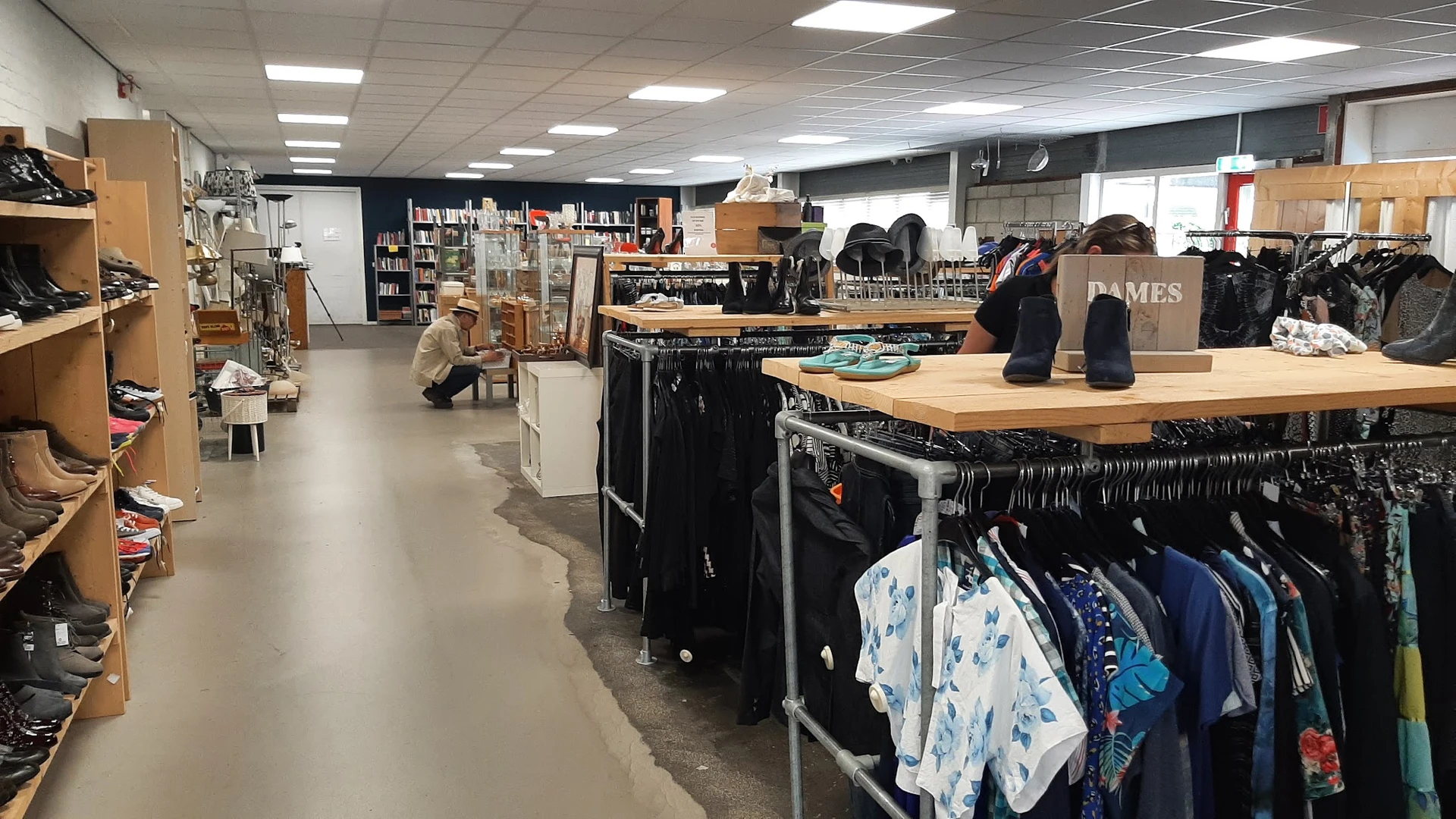 Binnenaanzicht van kringloopwinkel met kledingrekken, houten tafels en schappen met boeken en spullen