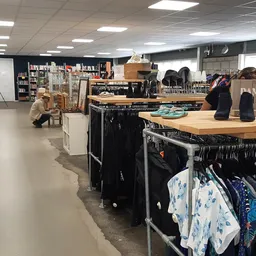 Binnenaanzicht van kringloopwinkel met kledingrekken, houten tafels en schappen met boeken en spullen