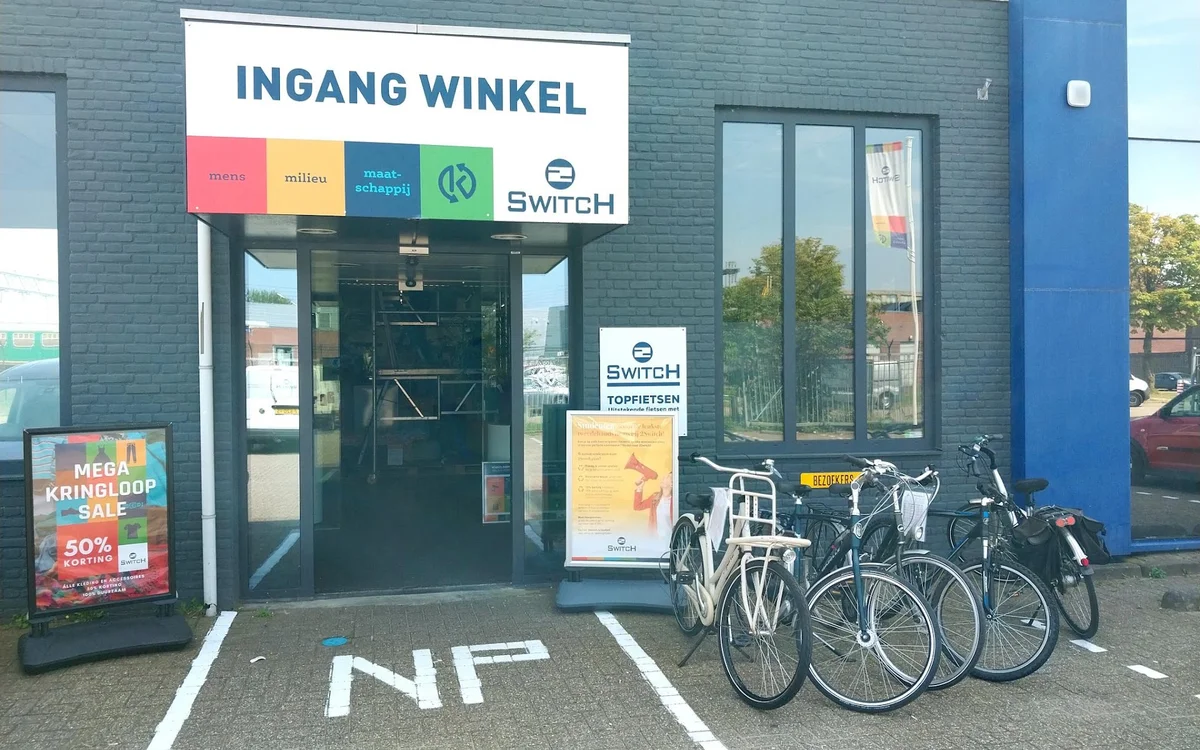 Winkelgevel met 'INGANG WINKEL' bord, merkenlogo's en fietsenstalling ervoor