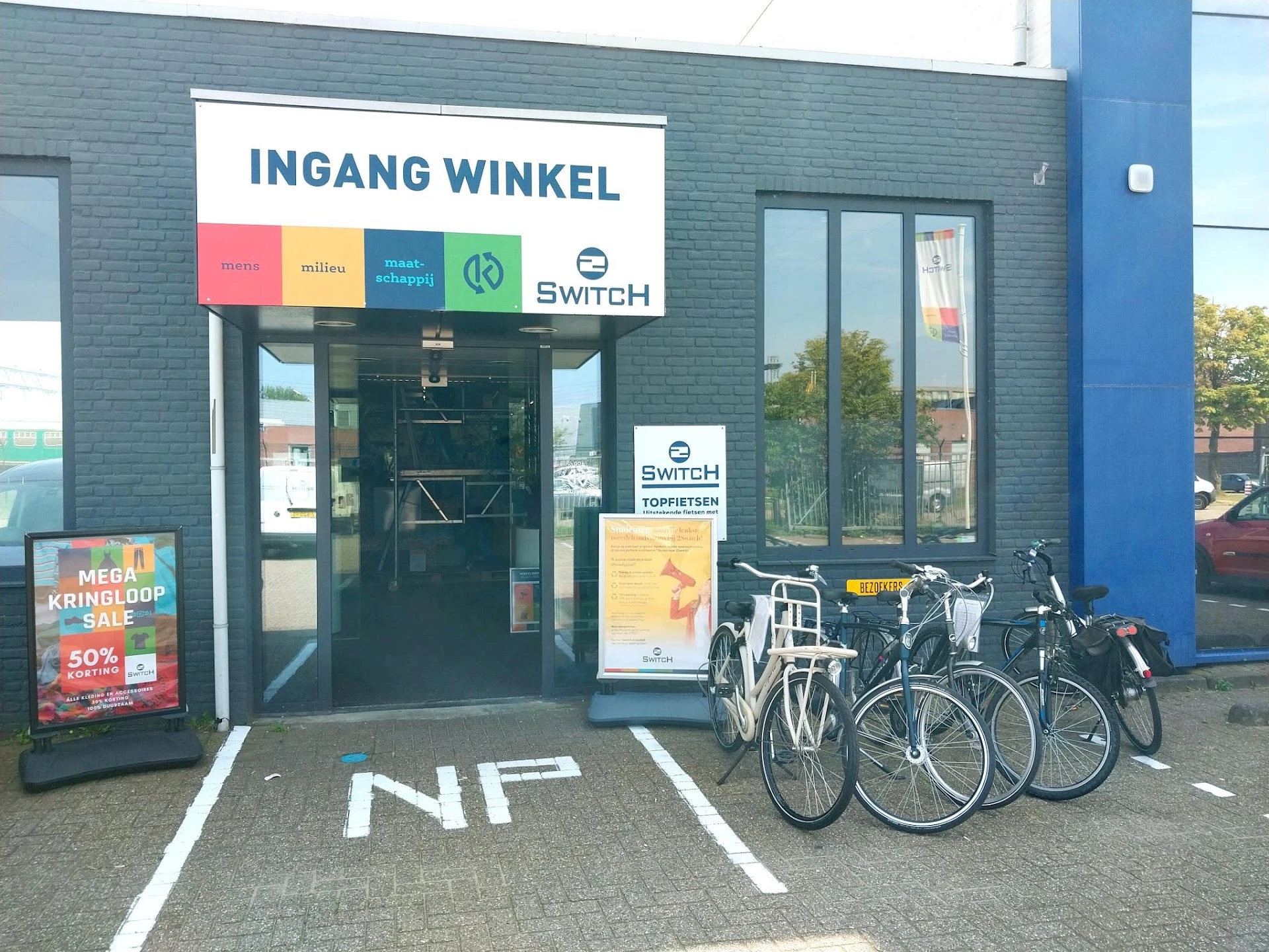 Winkelgevel met 'INGANG WINKEL' bord, merkenlogo's en fietsenstalling ervoor