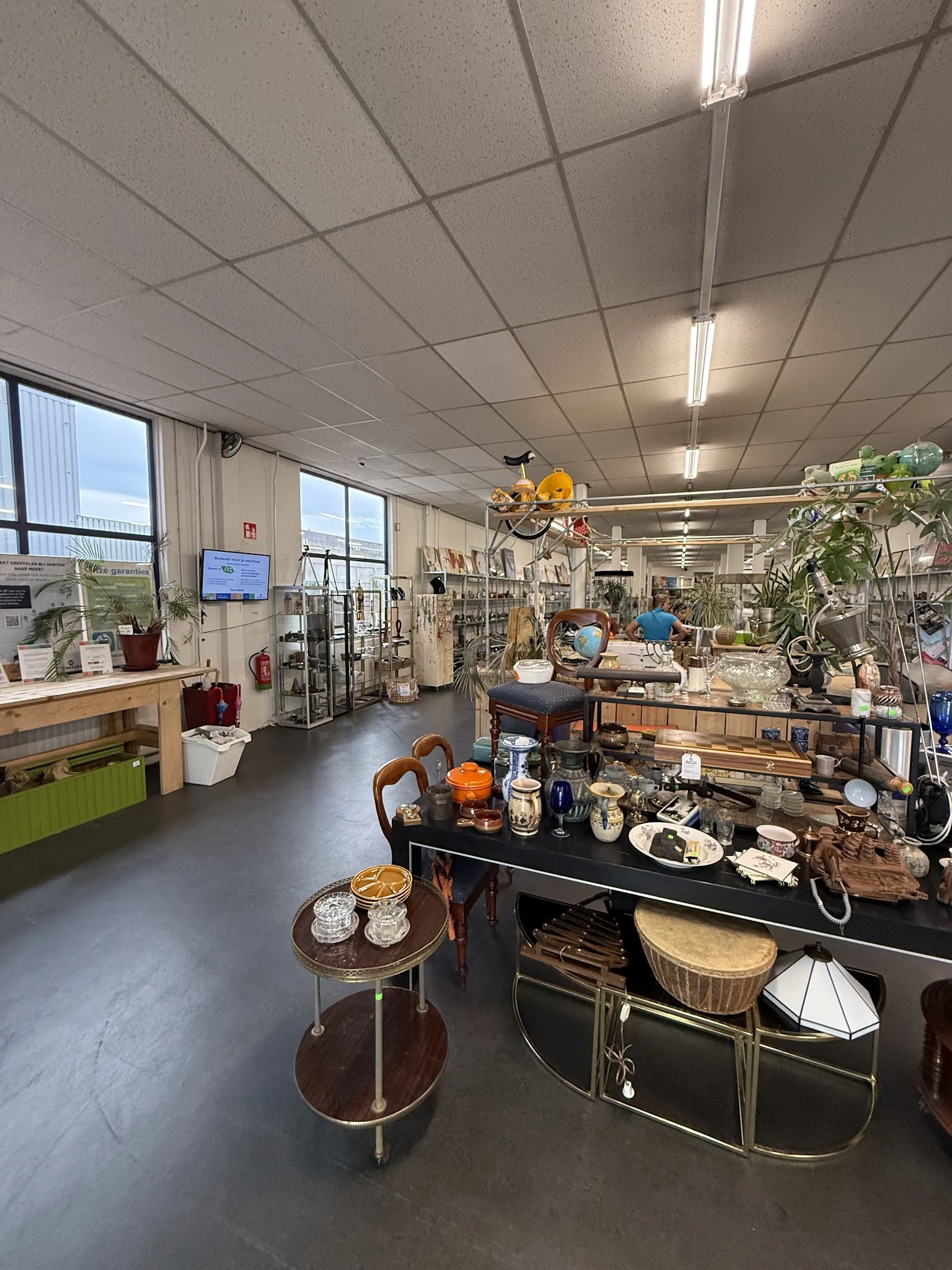 Ruime kringloopwinkel met diverse tweedehands spullen: meubels, huishoudartikelen, planten en decoratie uitgestald op tafels 