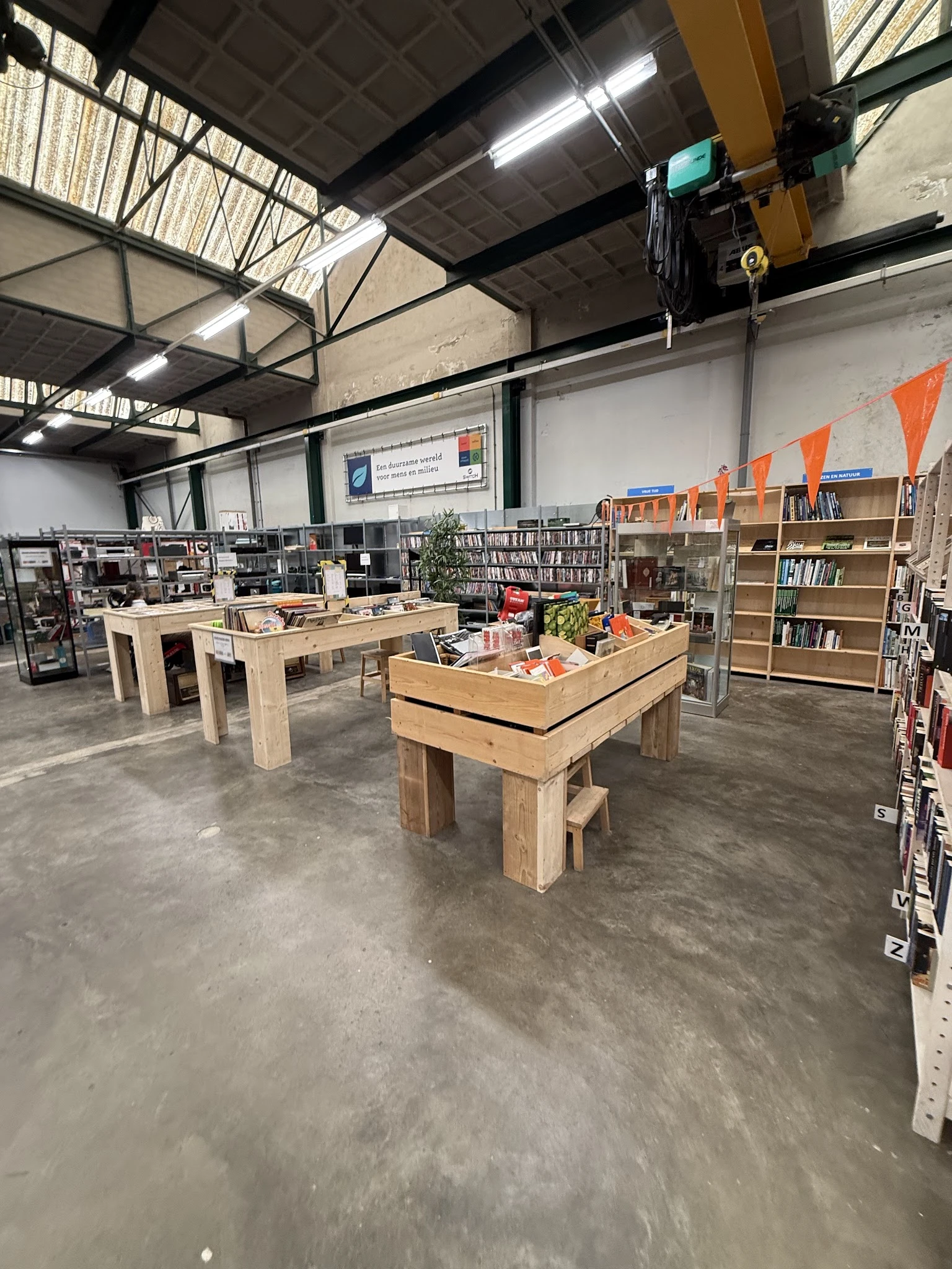 Winkelvloer met houten tafels, boekenkasten, stellingen en diverse tweedehands spullen in een ruime hal met industrieel plafo