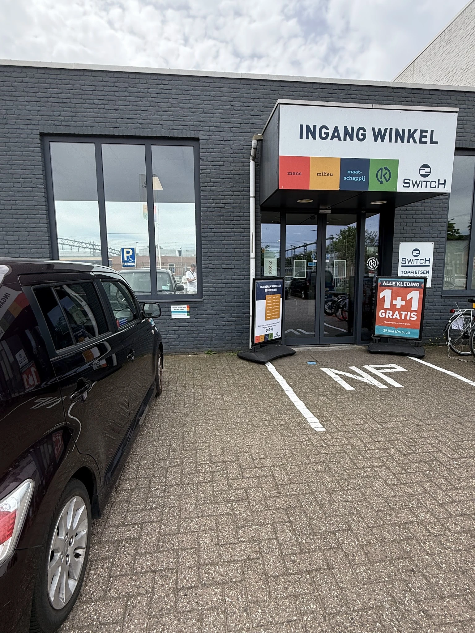Voorgevel van 2Switch kringloopwinkel met grijze bakstenen, glazen deuren en ramen, winkelnaam en logo zichtbaar, parkeerplaa
