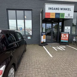 Voorgevel van 2Switch kringloopwinkel met grijze bakstenen, glazen deuren en ramen, winkelnaam en logo zichtbaar, parkeerplaa