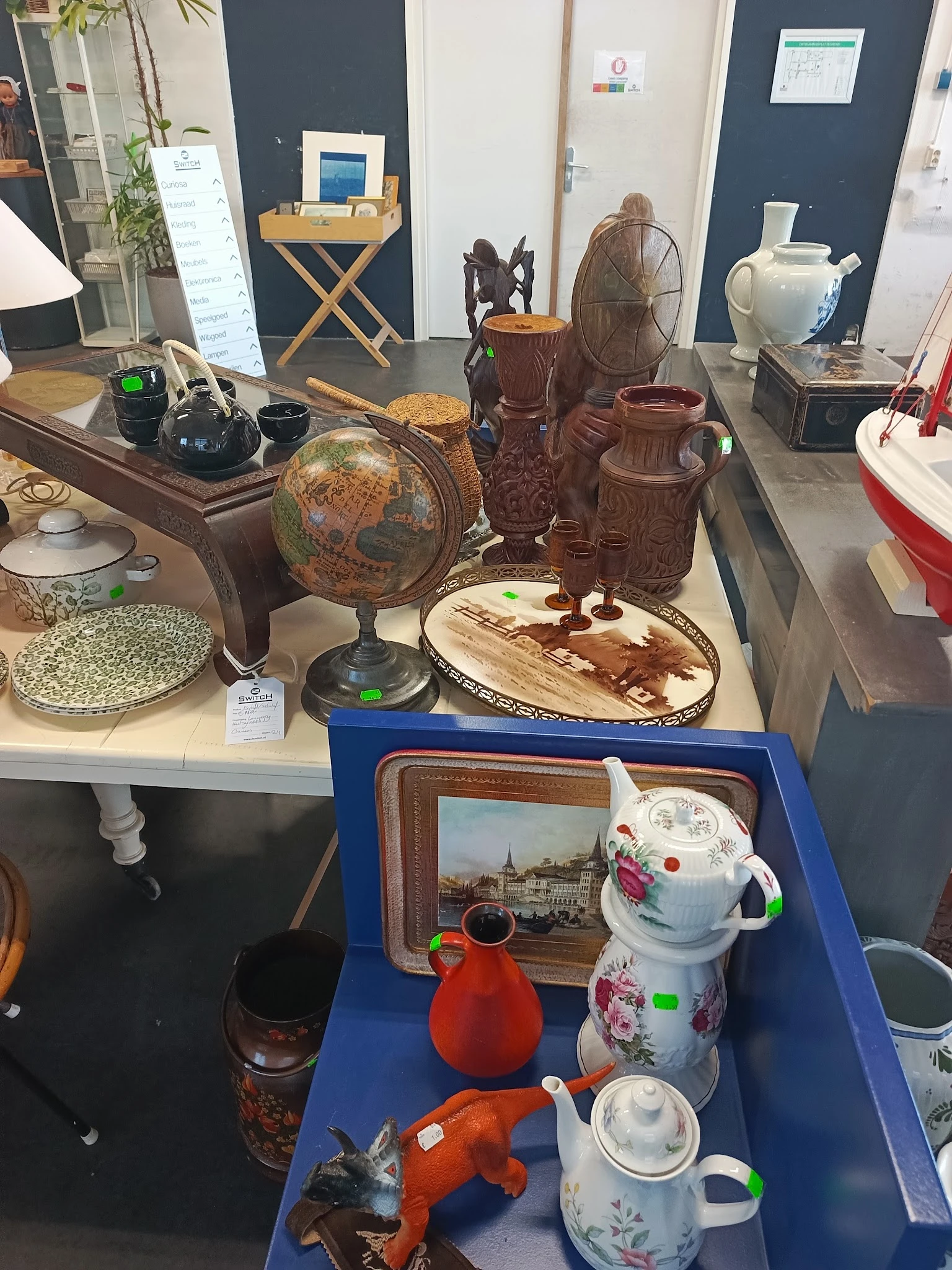 Uitgestalde tweedehandsartikelen op tafel: wereldbol, houten beelden, servies, decoratieve items en kunststofvoorwerpen