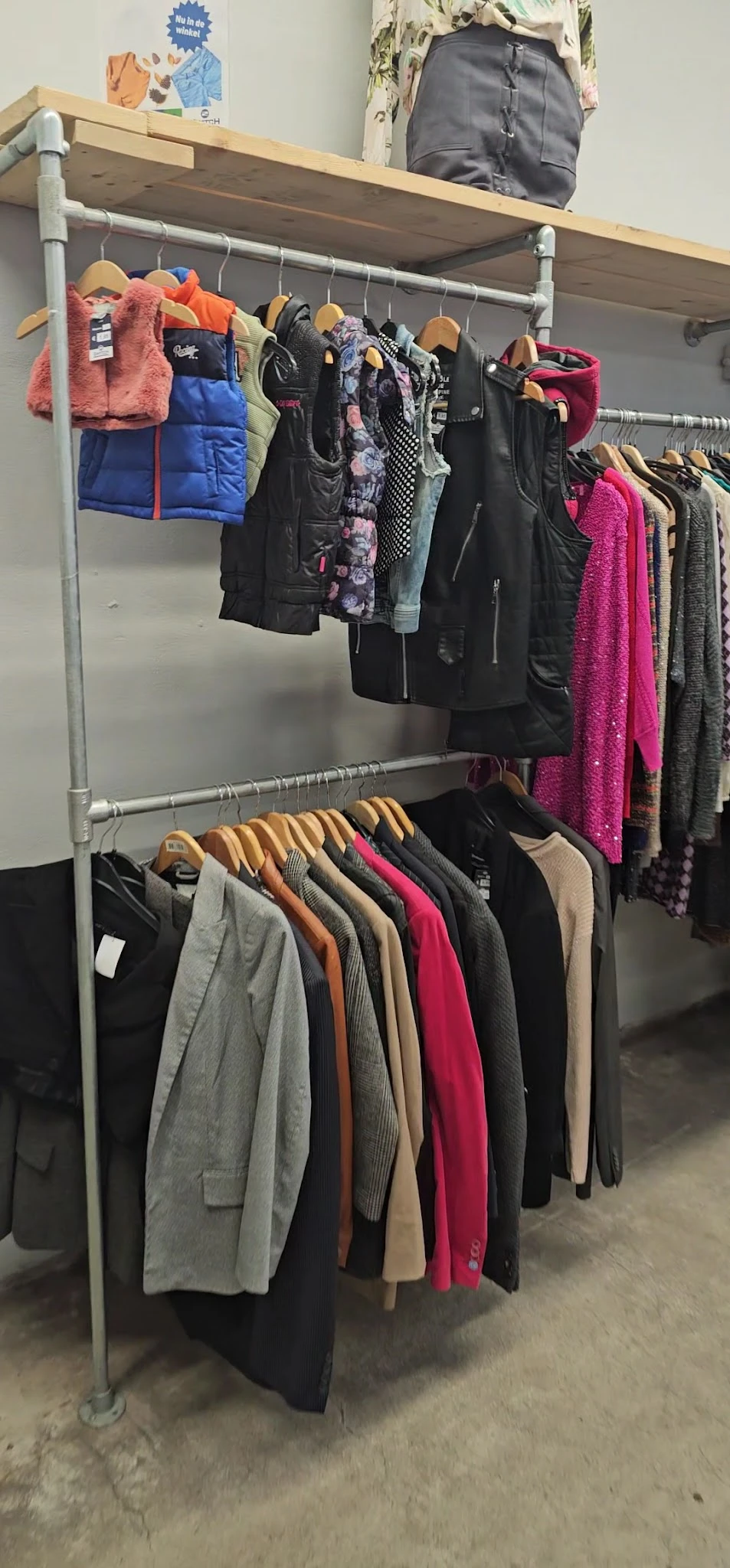 Rekken met tweedehands dameskleding op houten hangers, waaronder jassen, truien en jurken in verschillende kleuren