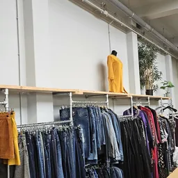Rekken met tweedehands kleding, waaronder jeans, donkere kleding en een geel kledingstuk op een plank erbovenop