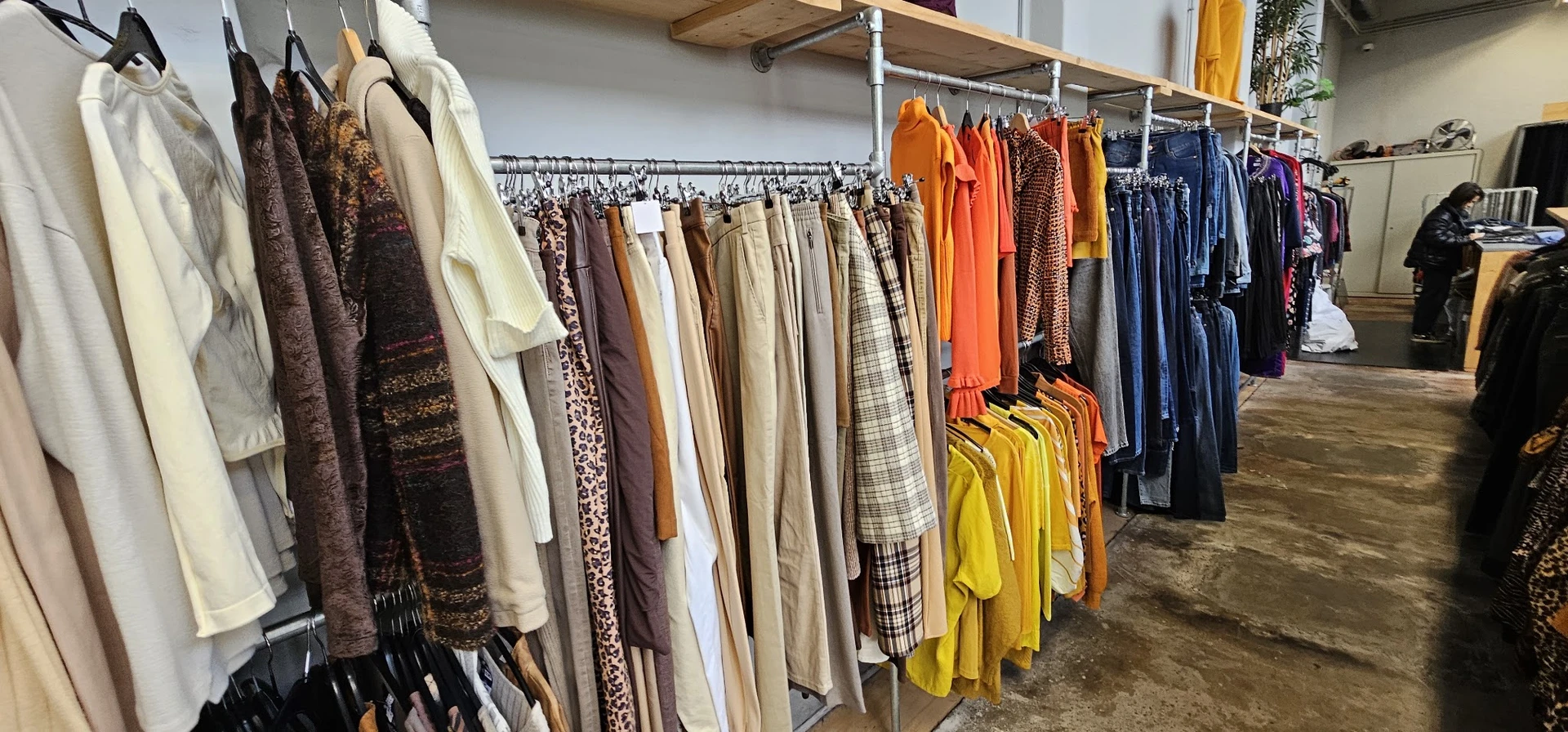 Rekken met tweedehands kleding in verschillende kleuren: bruinen, witte, beige, oranje, gele en blauwe kledingstukken op hang
