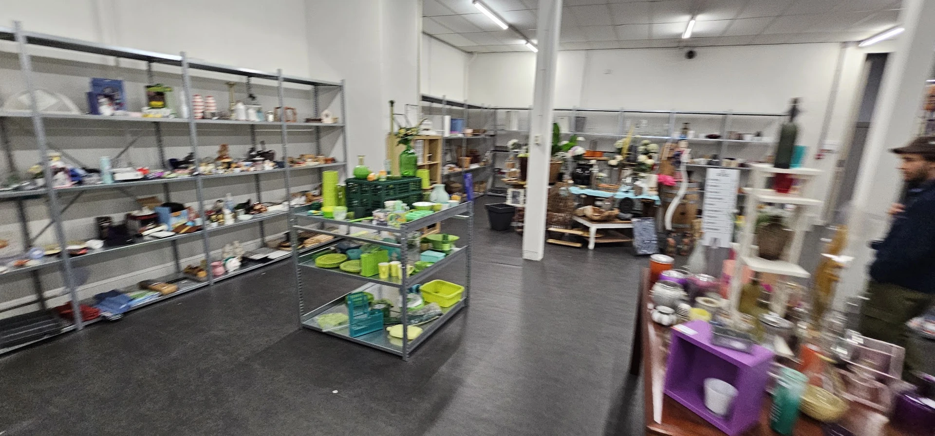 Winkelvloer van kringloopwinkel met diverse stellingen vol spullen, grijs beton, witte muren en kolommen