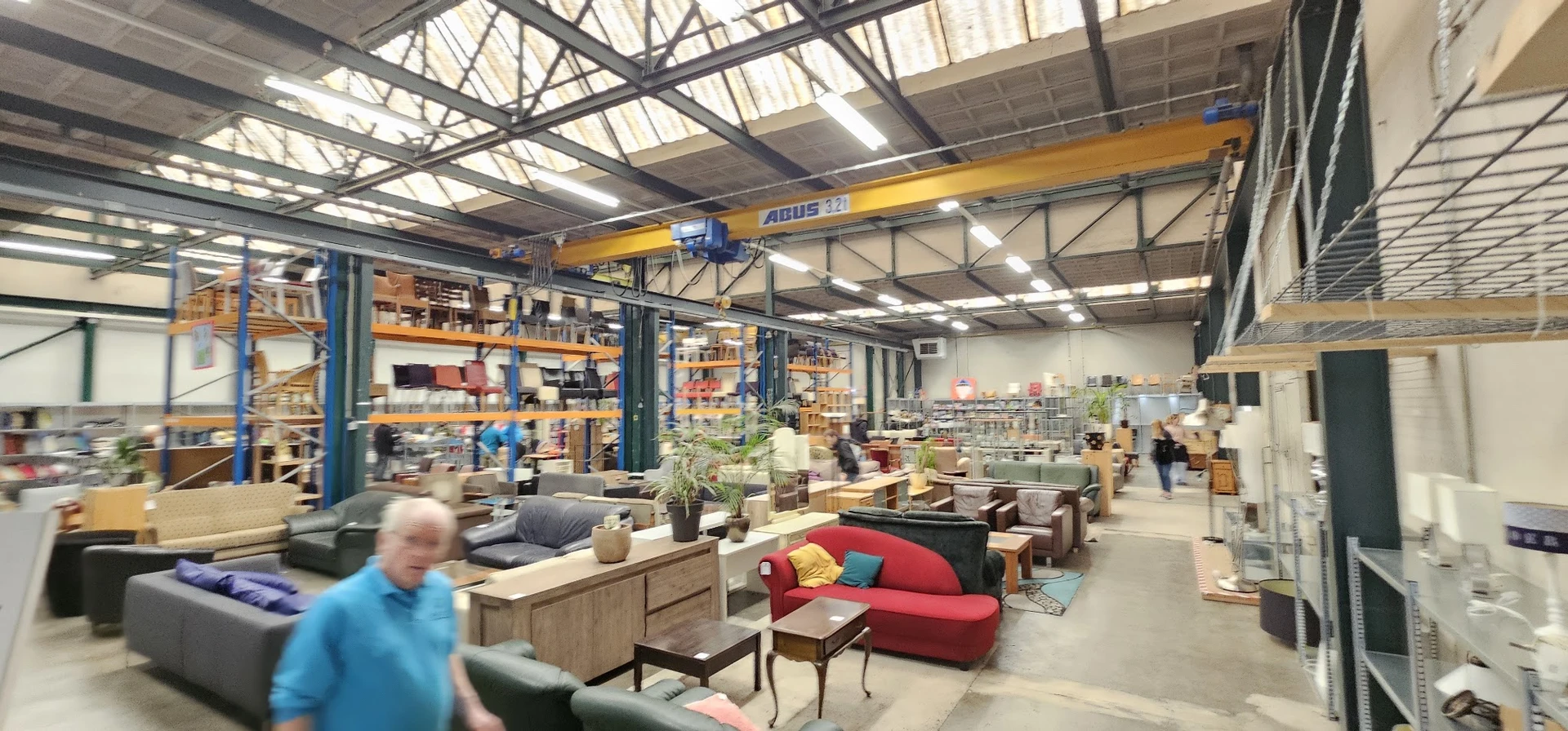 Ruime kringloopwinkel met diverse meubels, huishoudartikelen en spullen uitgestald op de vloer en stellingen onder een indust