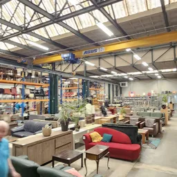 Ruime kringloopwinkel met diverse meubels, huishoudartikelen en spullen uitgestald op de vloer en stellingen onder een indust