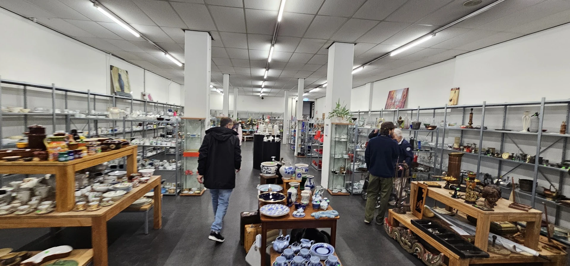 Winkelvloer met glazen vitrines, houten tafels met serviesgoed, stellingen en twee bezoekers in de kringloopwinkel