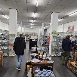 Winkelvloer met glazen vitrines, houten tafels met serviesgoed, stellingen en twee bezoekers in de kringloopwinkel