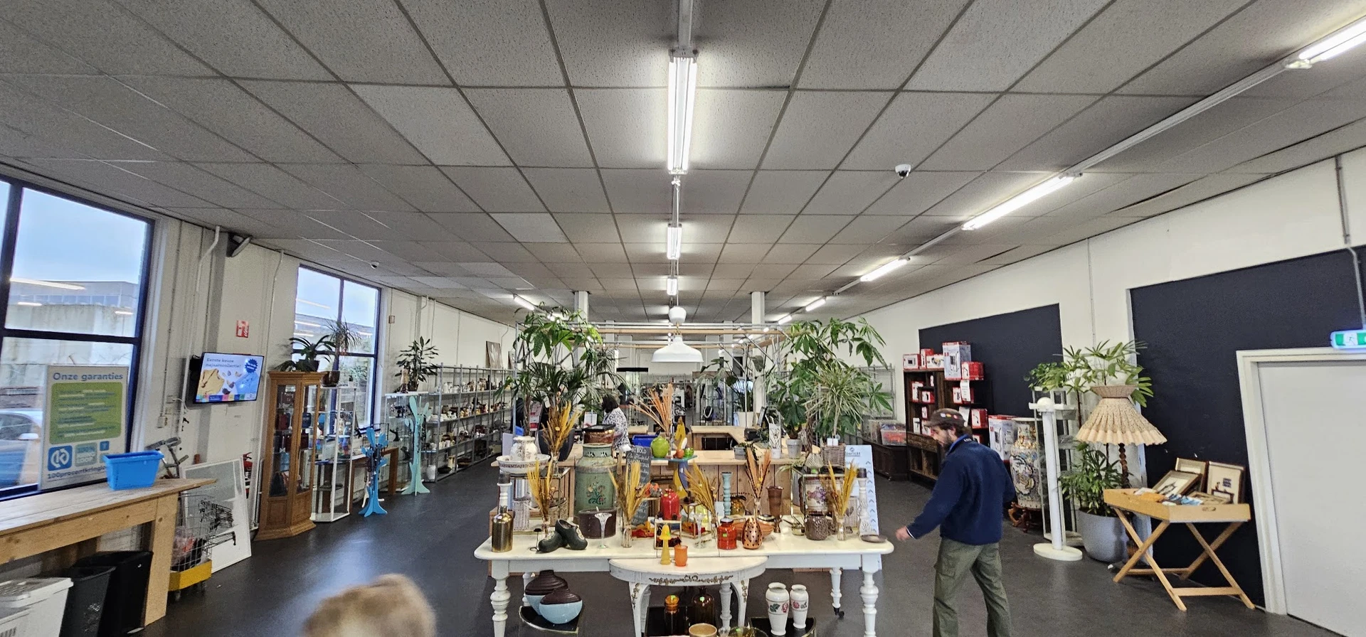 Overzicht van de winkelvloer met tafels vol huishoudartikelen, planten en decoratie, fluorescente verlichting aan plafond