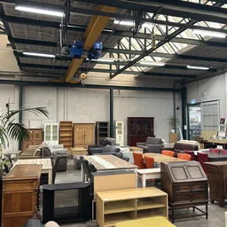 Ruim magazijn met tweedehands meubels, kasten, tafels en zitmeubelen uitgestald onder industrieel plafond met gele hijskraan