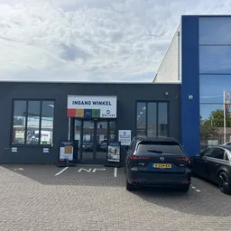 Zwarte gevel van kringloopwinkel met 'INGANG WINKEL' bordje en twee geparkeerde auto's op het terras