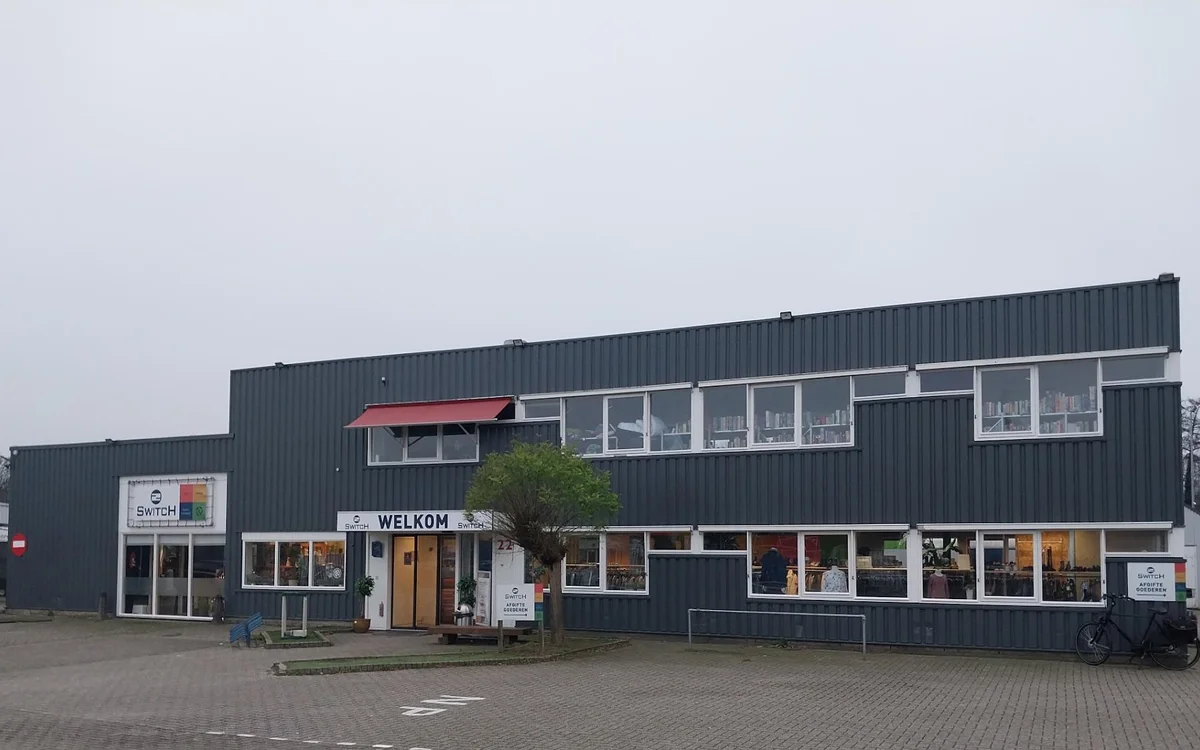 Voorgevel van kringloopwinkel 2Switch met donkergrijze golfplaten wanden, witte raampartijen en rode accenten, met 'WELKOM' o