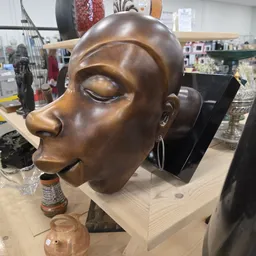 Bronzen decoratief hoofd met gesloten ogen en oorringen, geplaatst op een zwart sokkel in de winkel