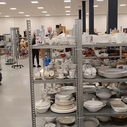Metalen stellingen volgestapeld met wit en grijs serviesgoed, borden, schalen en kopjes in een kringloopwinkel