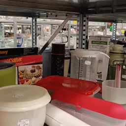 Diverse keukengerei en huishoudartikelen op een werktafel, waaronder rode schalen, een mixer, een snijder en andere gebruiksv