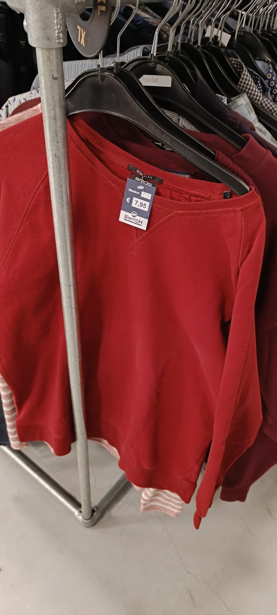 Close-up van een rood sweatshirt met label 2Switch, hangend aan een rek