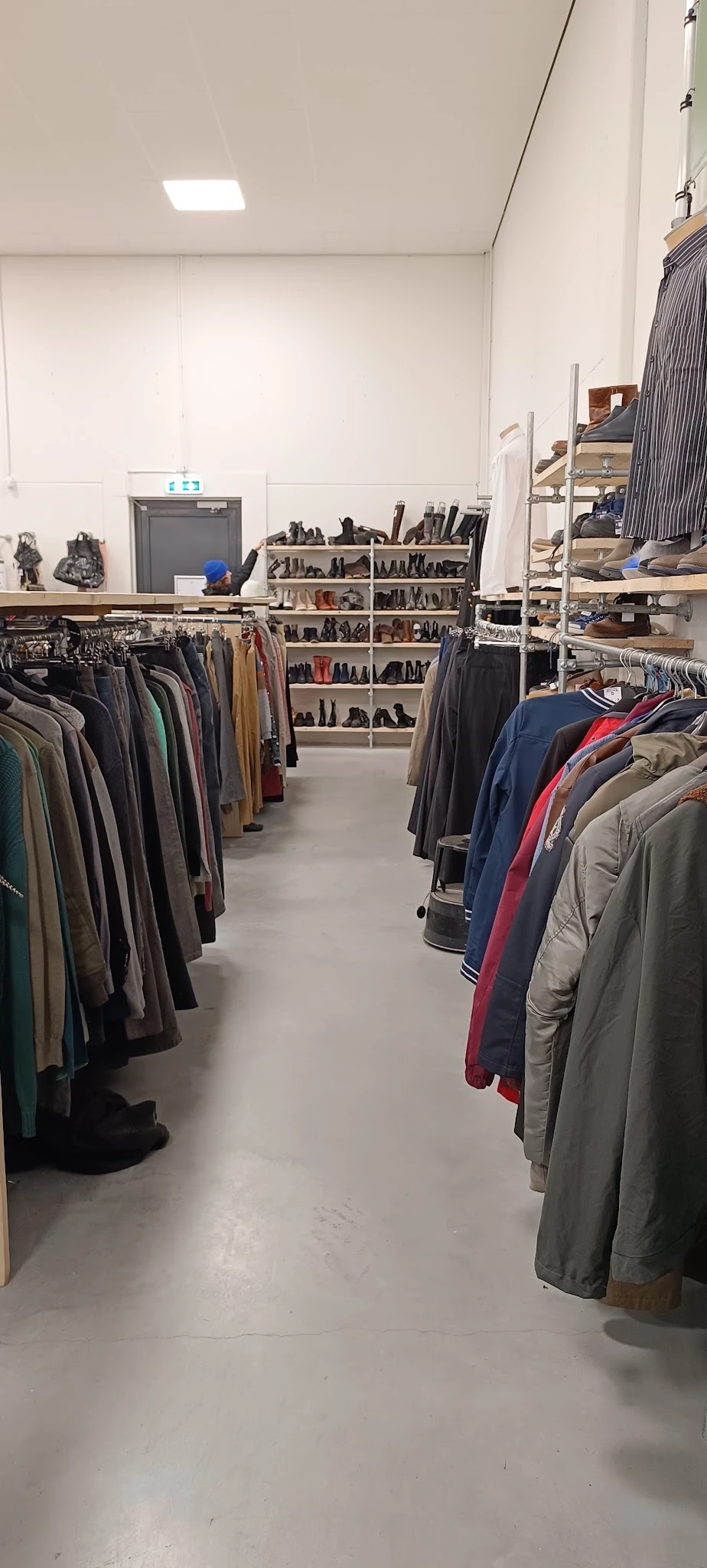 Rekken met tweedehands kleding, voornamelijk jassen en broeken, en stellingen met schoenen op de achtergrond