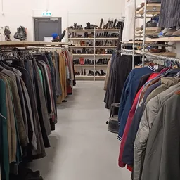 Rekken met tweedehands kleding, voornamelijk jassen en broeken, en stellingen met schoenen op de achtergrond