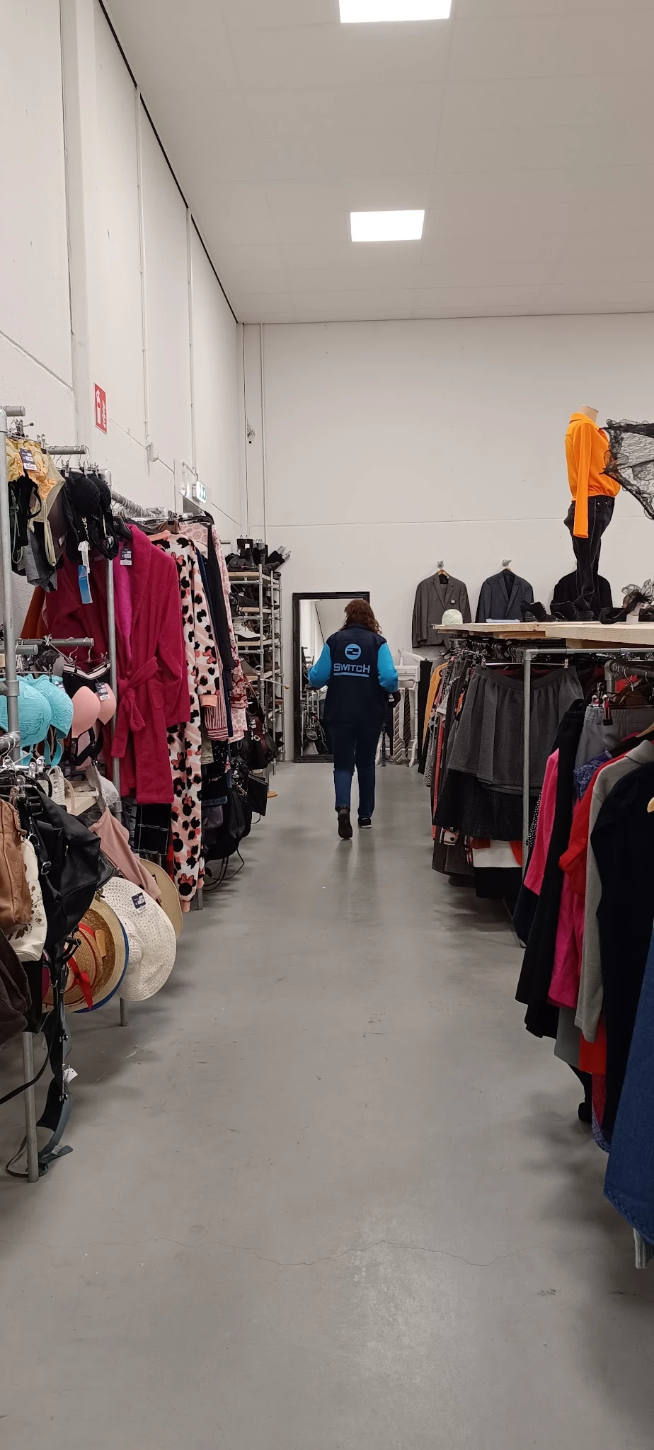 Winkelvloer van kringloopwinkel met rekken vol tweedehands kleding in verschillende kleuren, klant loopt door het gang