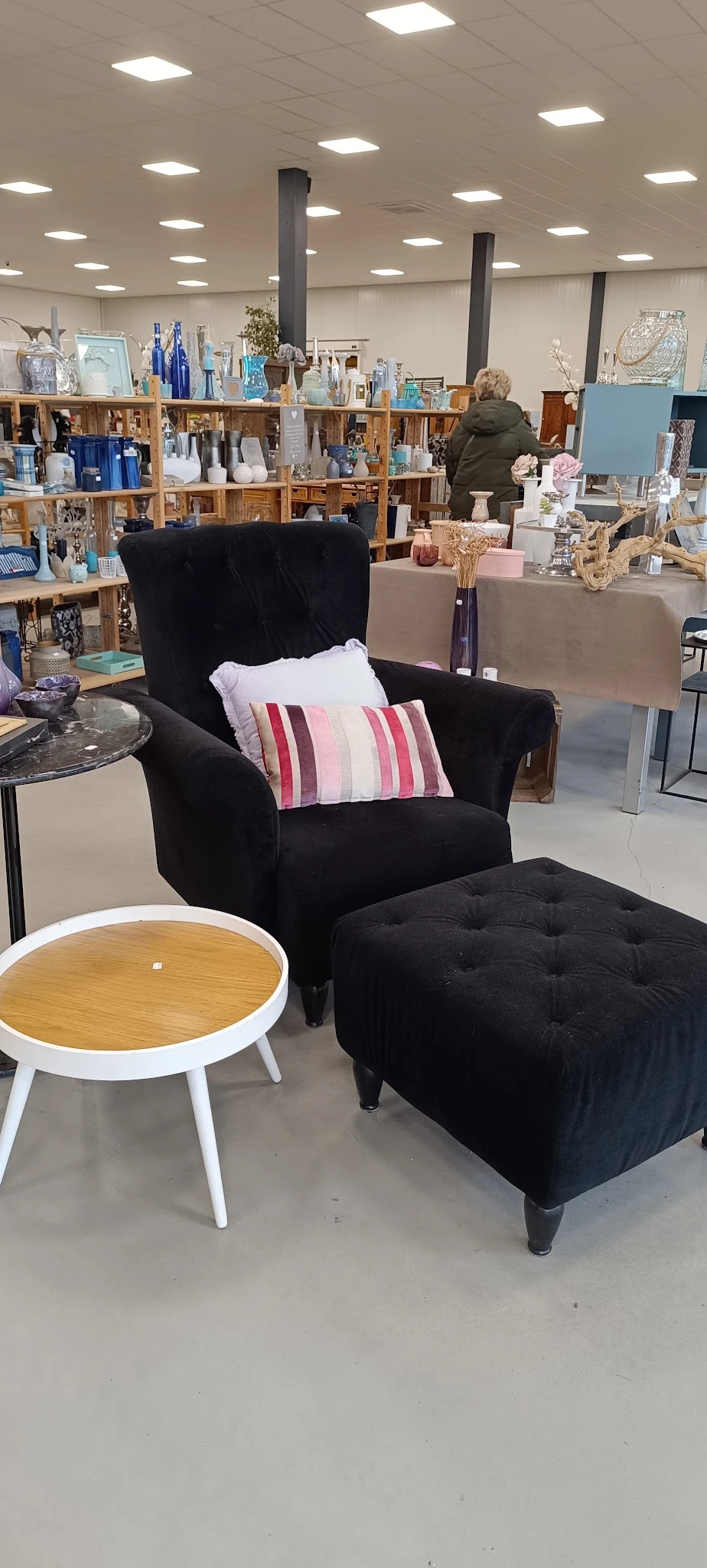 Zwarte fauteuil met kussens en zwarte pouf in een kringloopwinkel, met rond tafelblad op de achtergrond