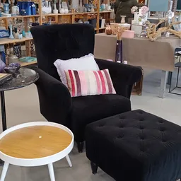 Zwarte fauteuil met kussens en zwarte pouf in een kringloopwinkel, met rond tafelblad op de achtergrond