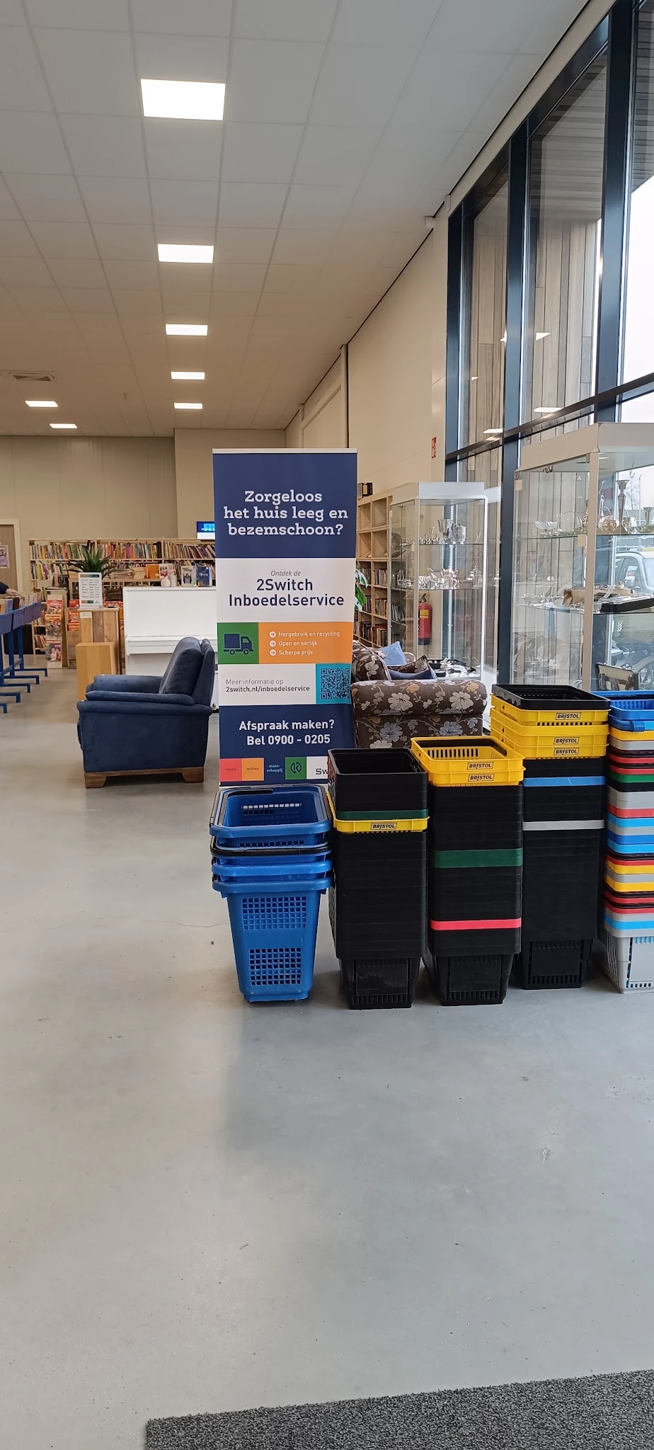 Winkelvloer met boekenkasten, een blauwe fauteuil, gekleurde kunststofkratten en service-informatieborden in een kringloopwin