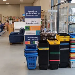 Winkelvloer met boekenkasten, een blauwe fauteuil, gekleurde kunststofkratten en service-informatieborden in een kringloopwin