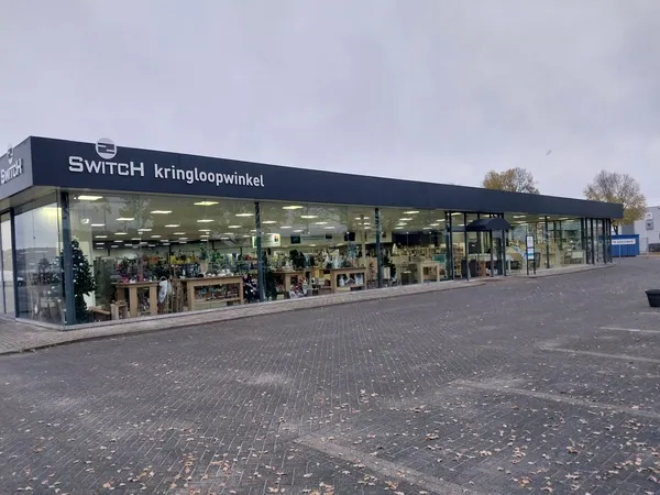 Winkelgevel van SWITCH kringloopwinkel met donkerblauwe luifel, grote ramen met goederen zichtbaar, op een plein met kasseien