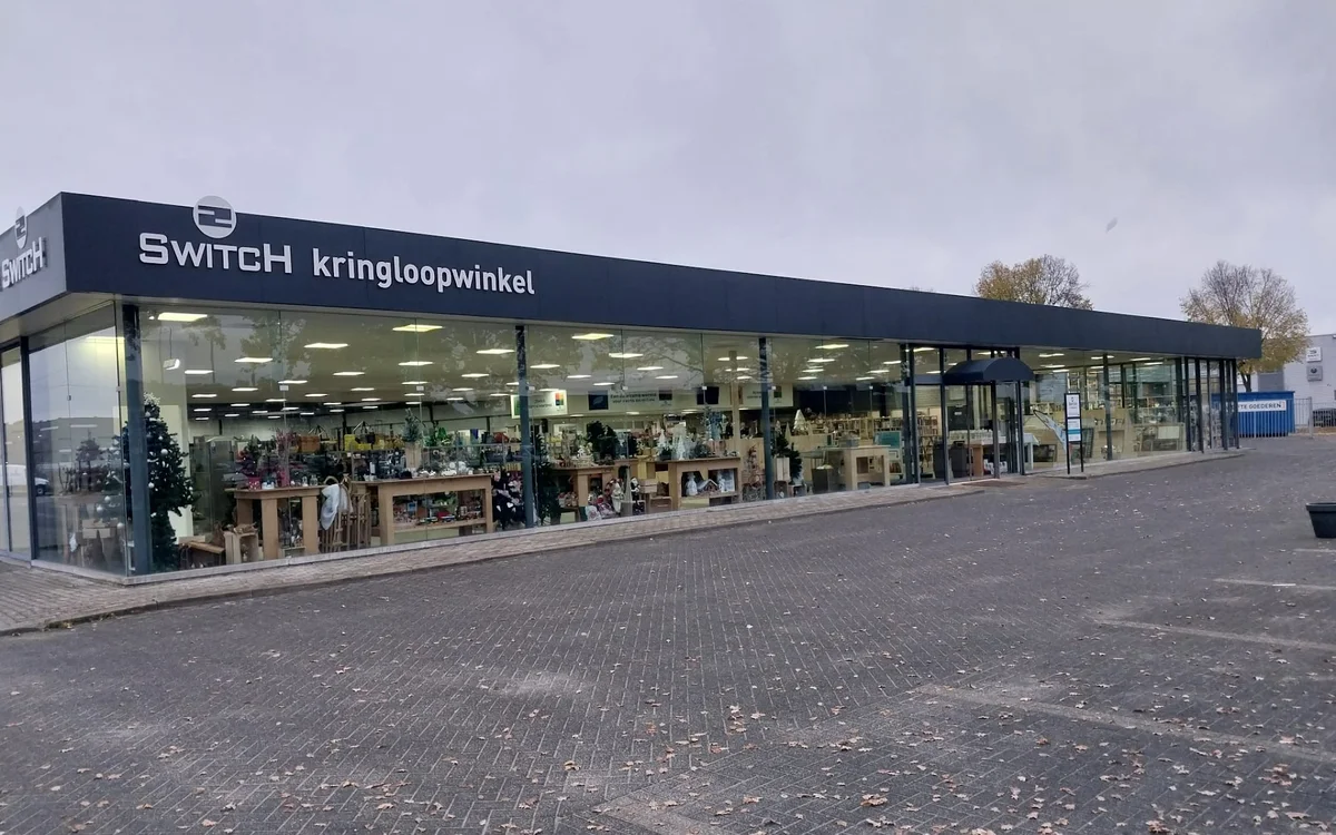 Winkelgevel van SWITCH kringloopwinkel met donkerblauwe luifel, grote ramen met goederen zichtbaar, op een plein met kasseien