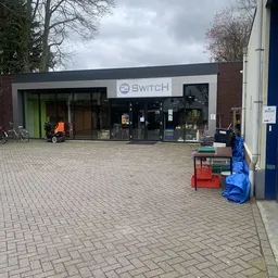 Voorgevel van kringloopwinkel 2Switch met groene en zwarte deuren, winkelnaam zichtbaar boven de ingang
