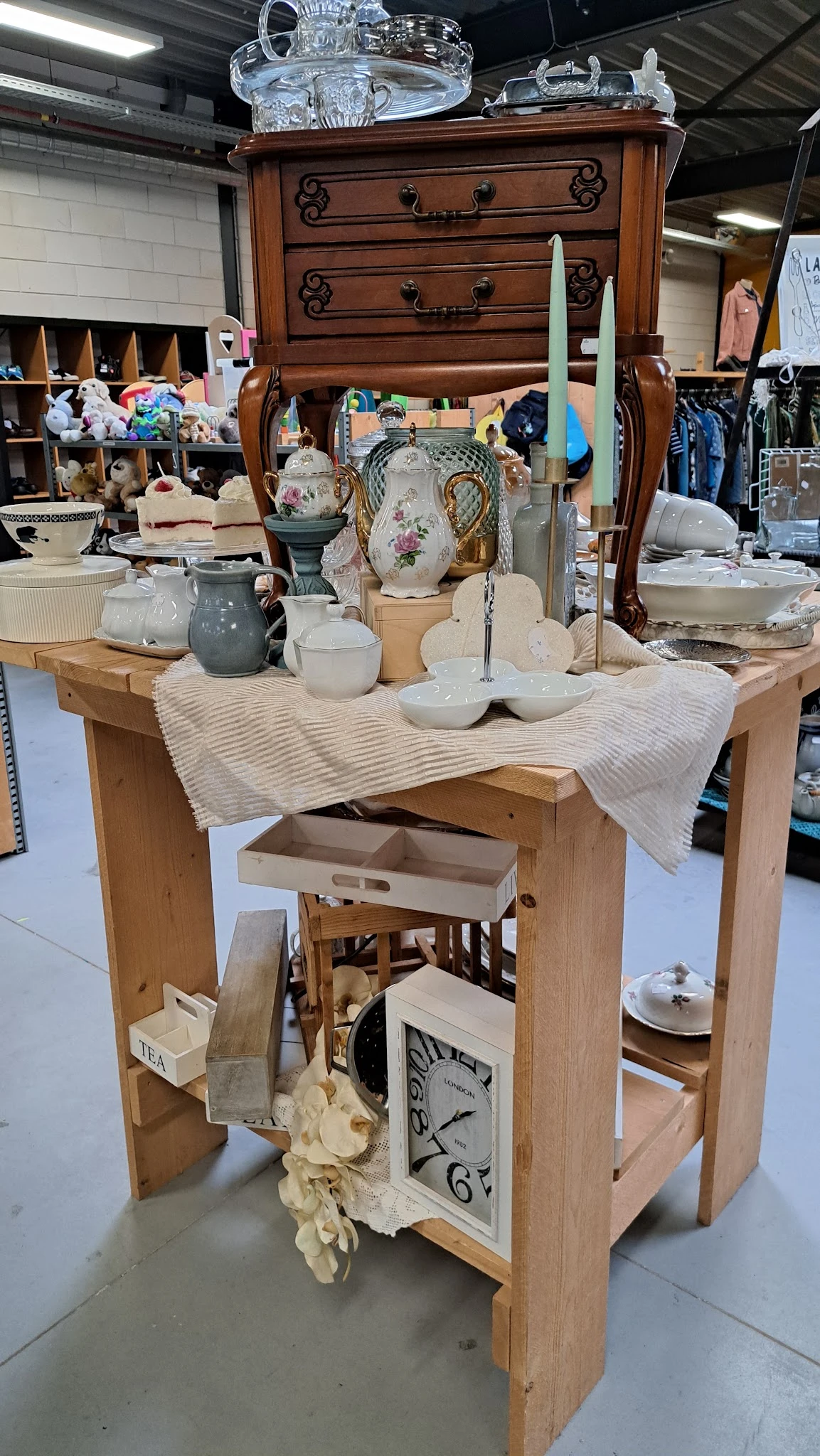 Houten tafel met wit serviesgoed, kandelaars en decoratieve items uitgestald in een kringloopwinkel