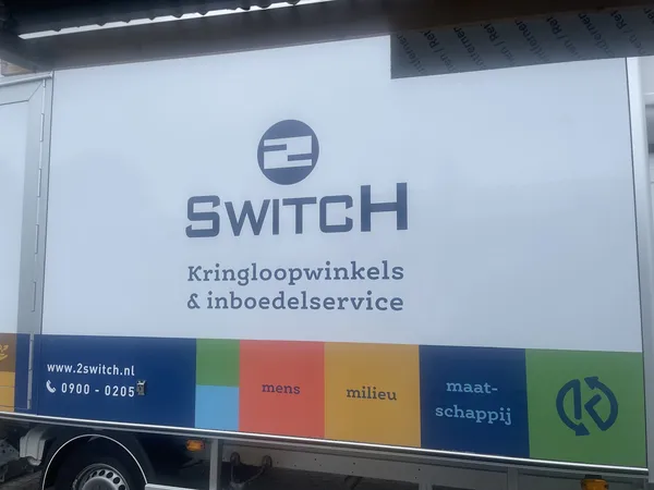 Witte gevel van kringloopwinkel 2Switch met logo en tekst, donkerblauwe band met gekleurde vakken onderaan