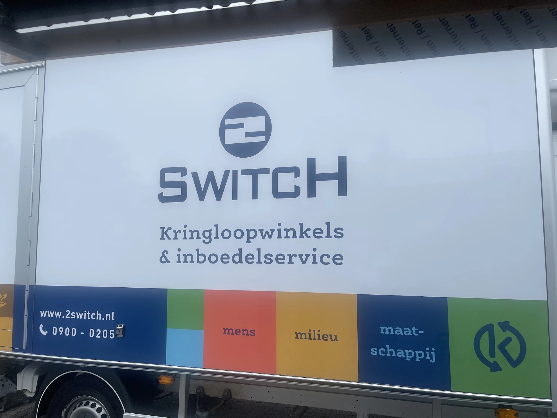 Witte gevel van kringloopwinkel 2Switch met logo en tekst, donkerblauwe band met gekleurde vakken onderaan