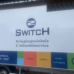 Witte gevel van kringloopwinkel 2Switch met logo en tekst, donkerblauwe band met gekleurde vakken onderaan