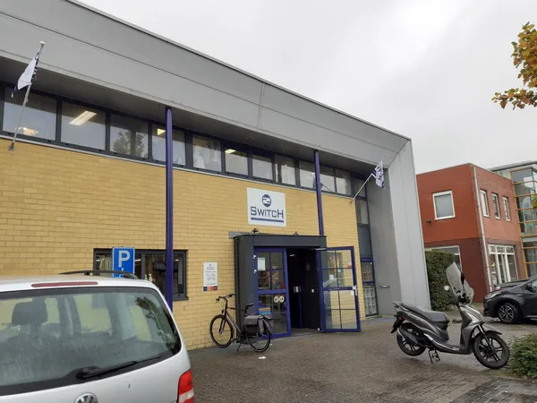 Geel en grijs gevelfront van kringloopwinkel 2Switch met blauwe deuren, parkeerplaats en fietsen vooraan.