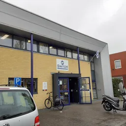 Geel en grijs gevelfront van kringloopwinkel 2Switch met blauwe deuren, parkeerplaats en fietsen vooraan.