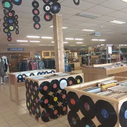 Uitgestalde vinyl platen in houten bakken in de winkel, met op de achtergrond kledingrekken en andere tweedehands items