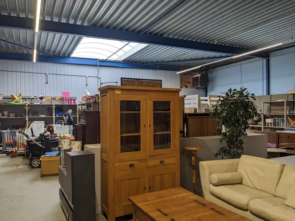 Winkelvloer met meubels: houten vitrinekast, houten tafel en crèmekleurige bank, omgeven door diverse tweedehands artikelen e