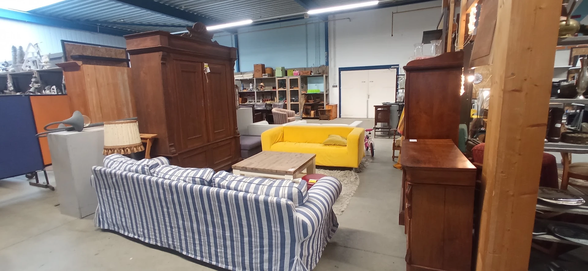 Winkelvloer met diverse meubels: gele bank, gestreepte fauteuil, houten kasten en stellingen met spullen op de achtergrond