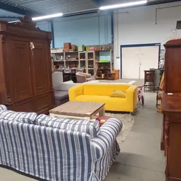 Winkelvloer met diverse meubels: gele bank, gestreepte fauteuil, houten kasten en stellingen met spullen op de achtergrond