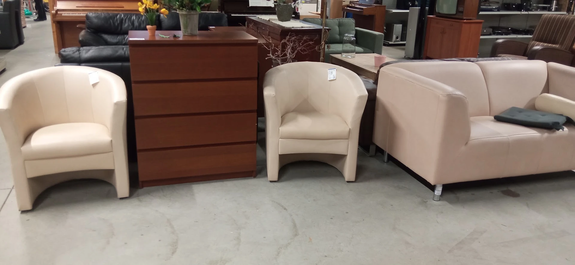 Uitgestalde fauteuils en dressoir in een kringloopwinkel, waaronder een beige en roze stoelen en een bruine houten kast