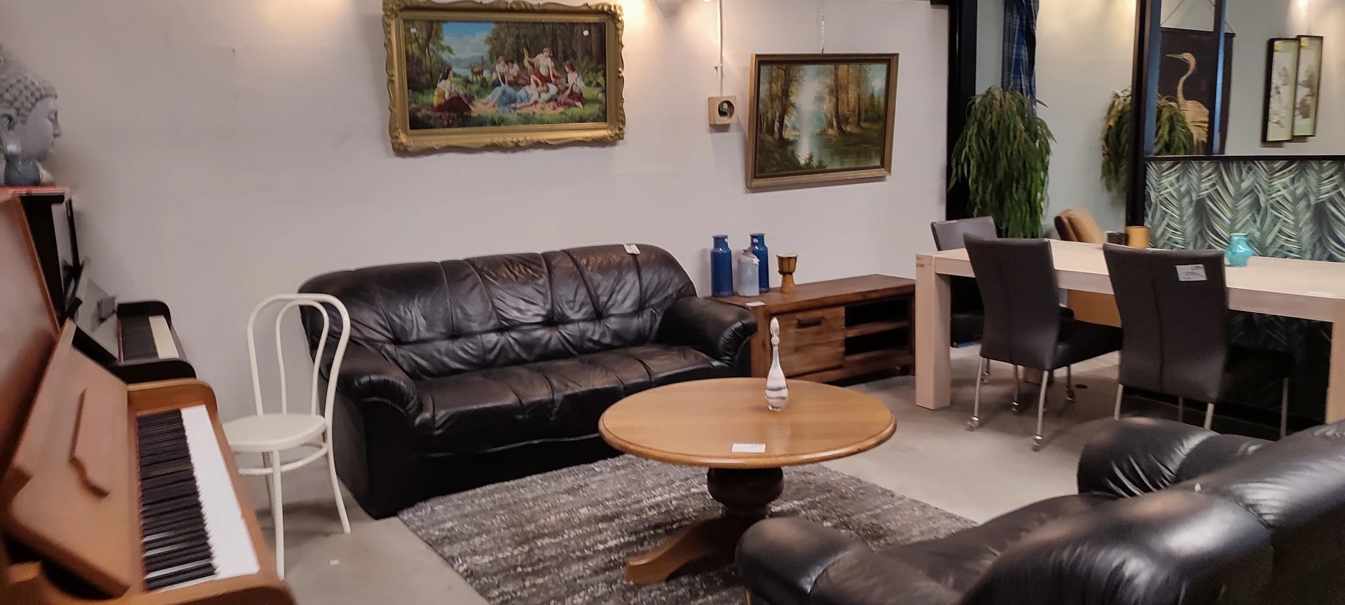 Woonkamer met zwarte leren bank, ronde houten tafel, schilderijen aan muur en verschillende meubelstukken