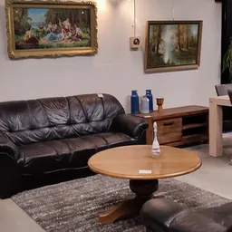 Woonkamer met zwarte leren bank, ronde houten tafel, schilderijen aan muur en verschillende meubelstukken